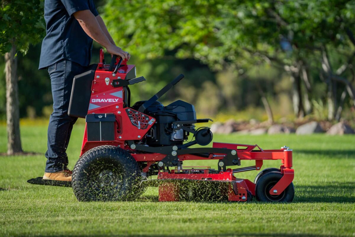 Person operating a red Gravely stand-on lawnmower, cutting grass in a landscaped area with trees in the background.