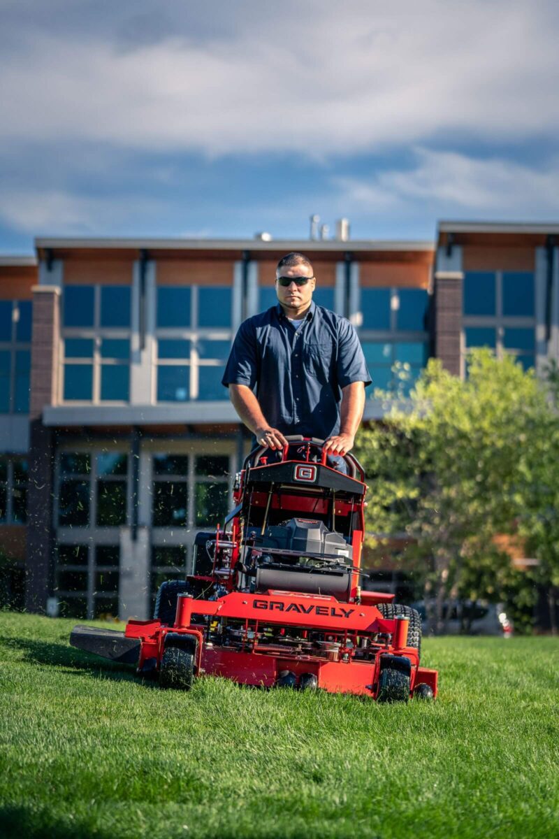 A person operates a red Gravely lawn mower on a grassy area in front of a modern building on a sunny day.