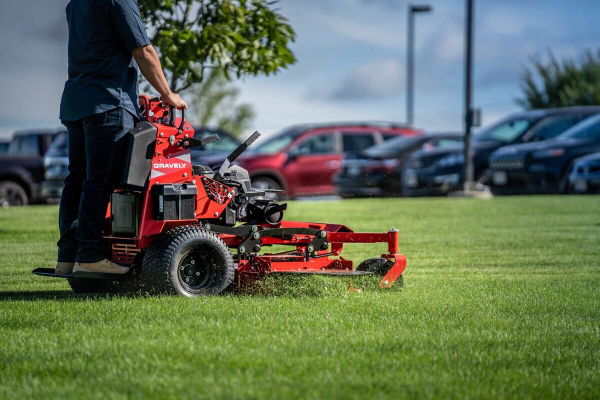 Person operating a red Gravely lawn mower on a grassy area, with parked cars and trees in the background.