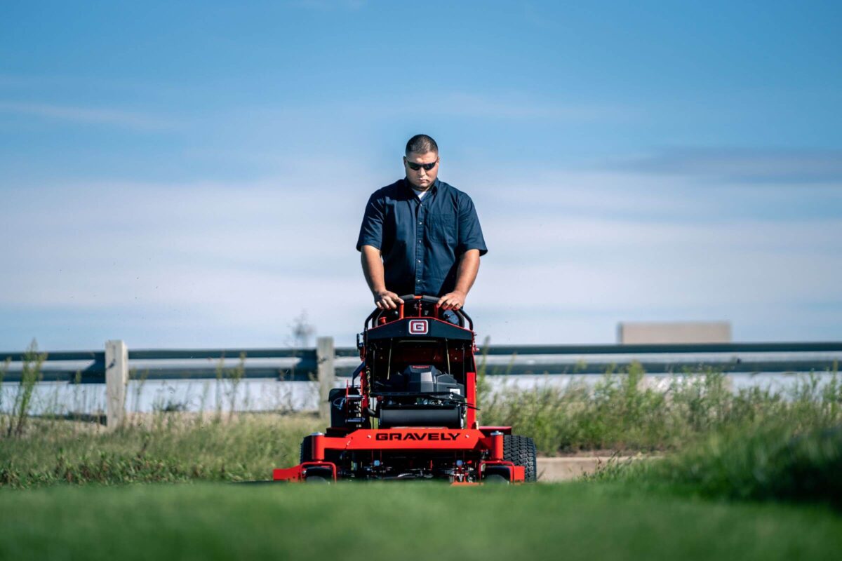 A person operates a red Gravely lawn mower on a grass field under a clear blue sky.