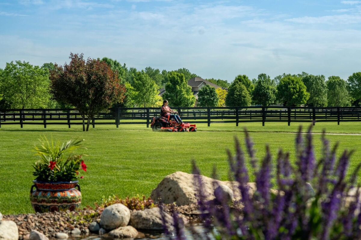 Person mowing a large, green lawn on a riding mower. The lawn is bordered by a black wooden fence and surrounded by trees. The foreground has flowers and rocks near a pond.