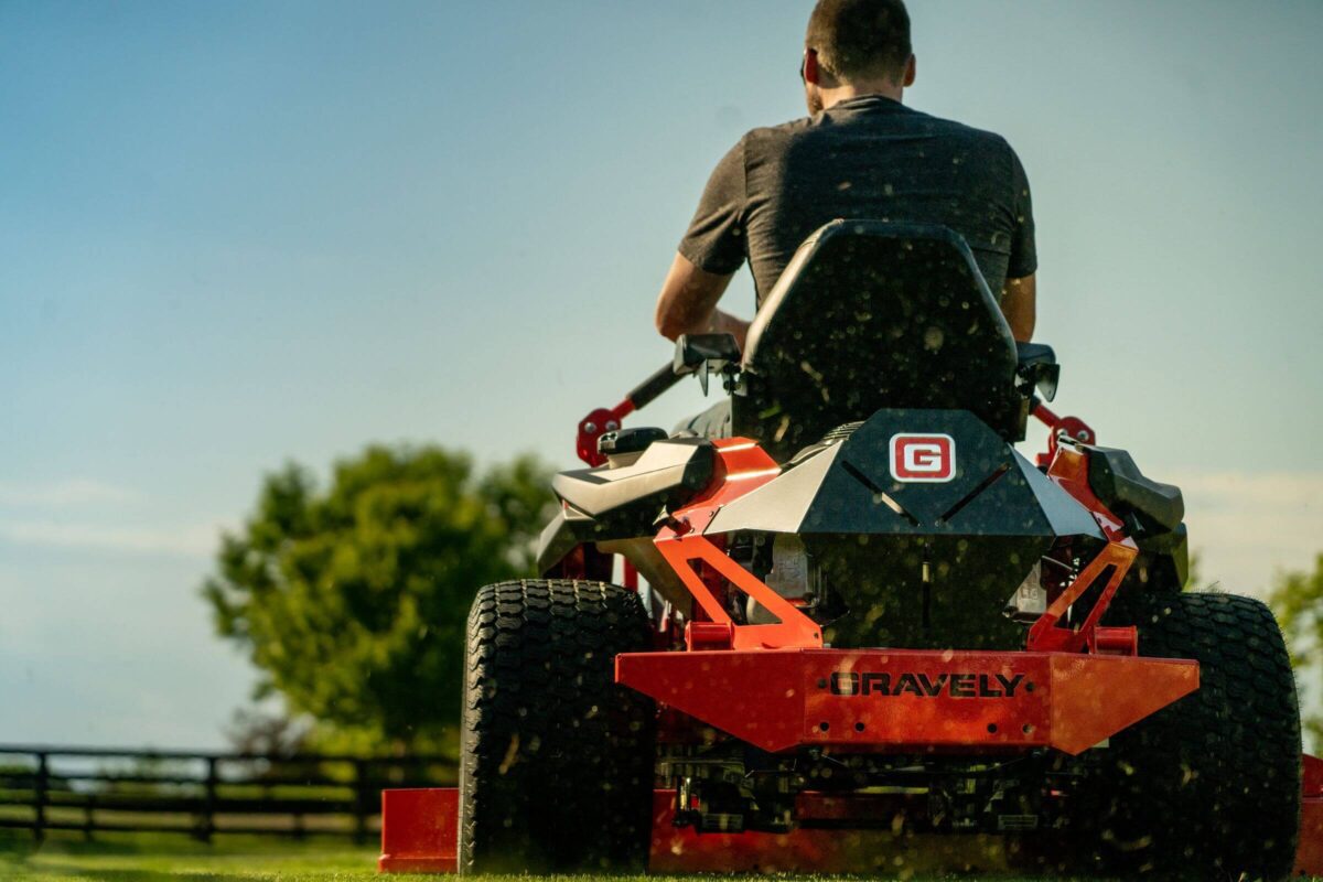 A person operates an orange Gravely lawn mower on grassy terrain, with trees and a fence in the background.