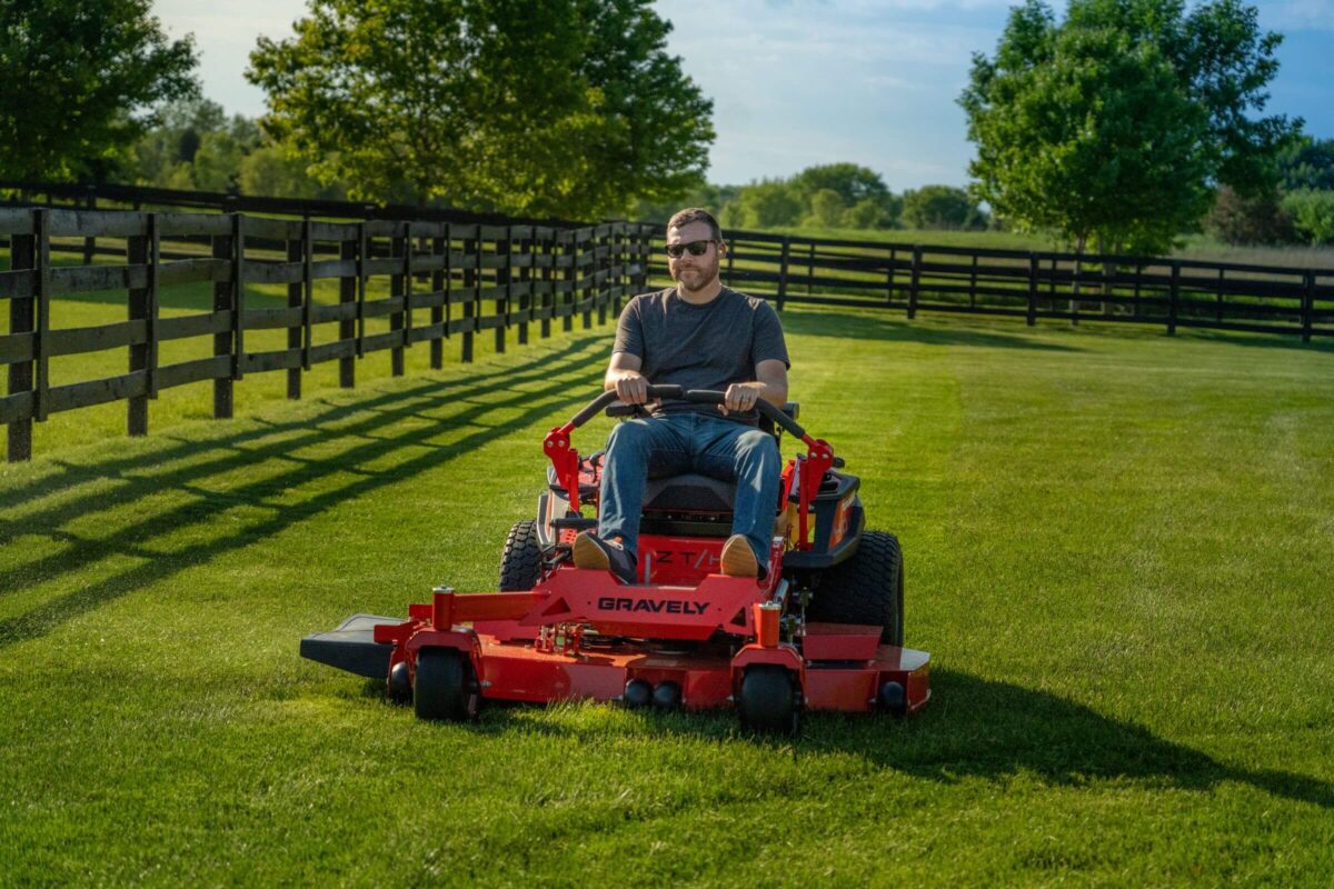 A person operates a Gravely lawn mower on a grassy field, with wooden fences and trees in the background under a blue sky.