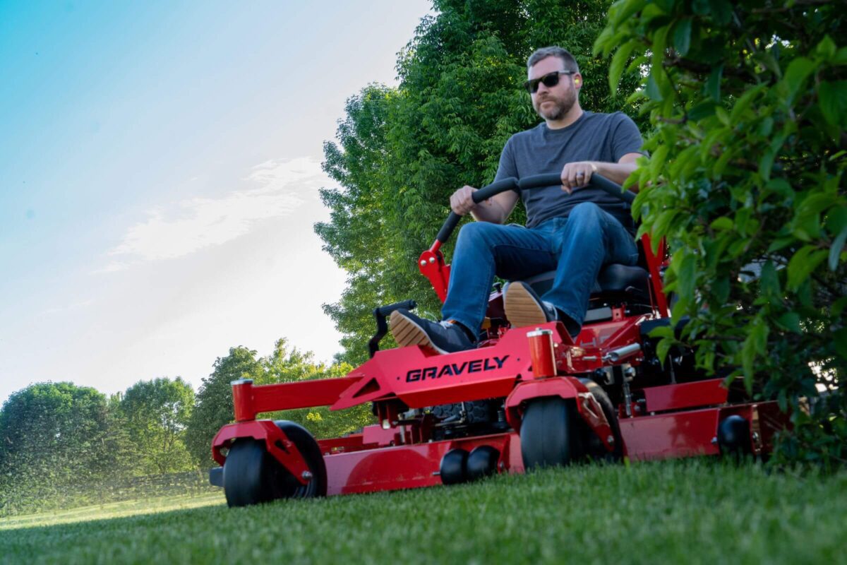 A man wearing sunglasses is mowing the lawn with a red Gravely ride-on lawnmower.