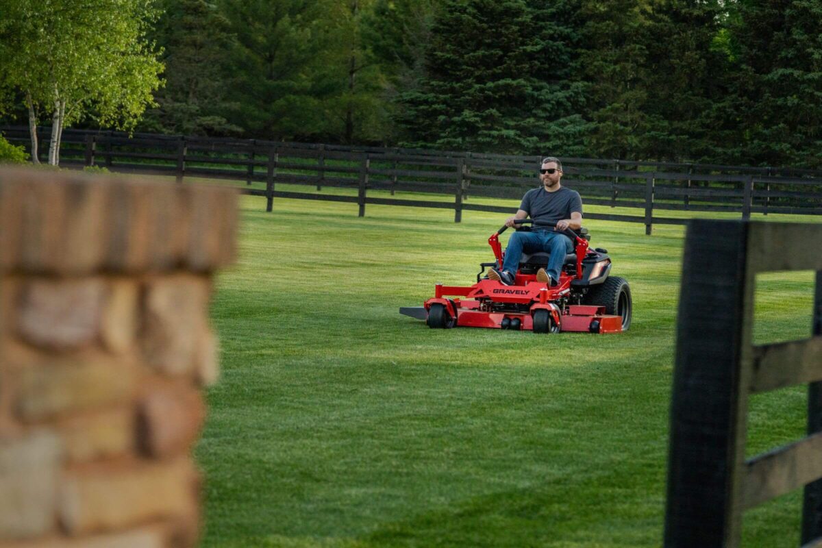 A person is riding a red lawn mower on a neatly trimmed grass lawn, surrounded by wooden fencing and trees in the background.