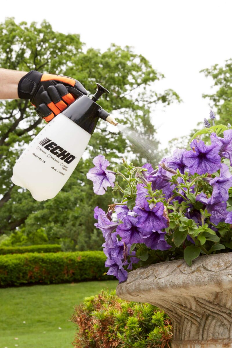 A person wearing gloves sprays purple flowers in a stone planter with a handheld sprayer.