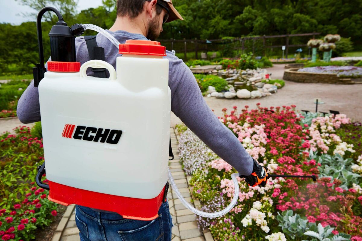 A person wearing a cap uses a backpack sprayer to water flowers in a garden.