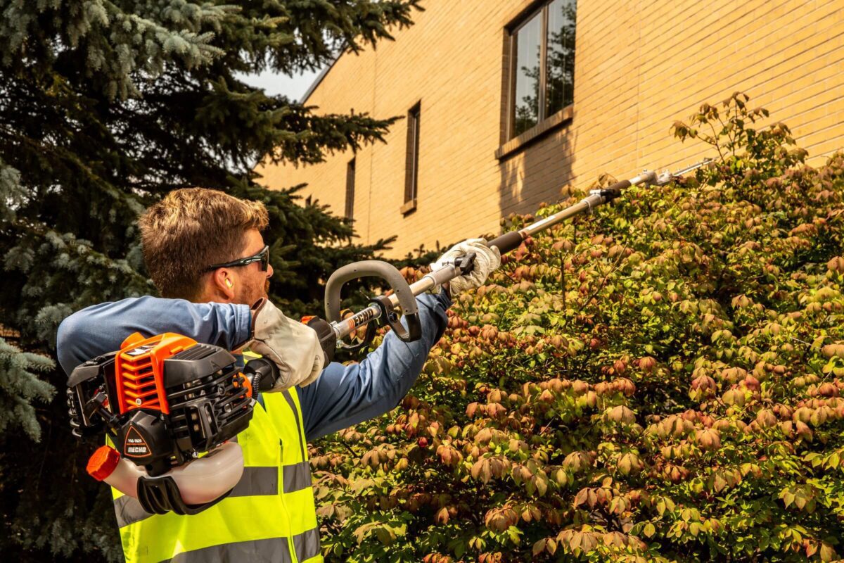 Person in a high-visibility vest uses a long-handled hedge trimmer to trim bushes near a building, with trees in the background.