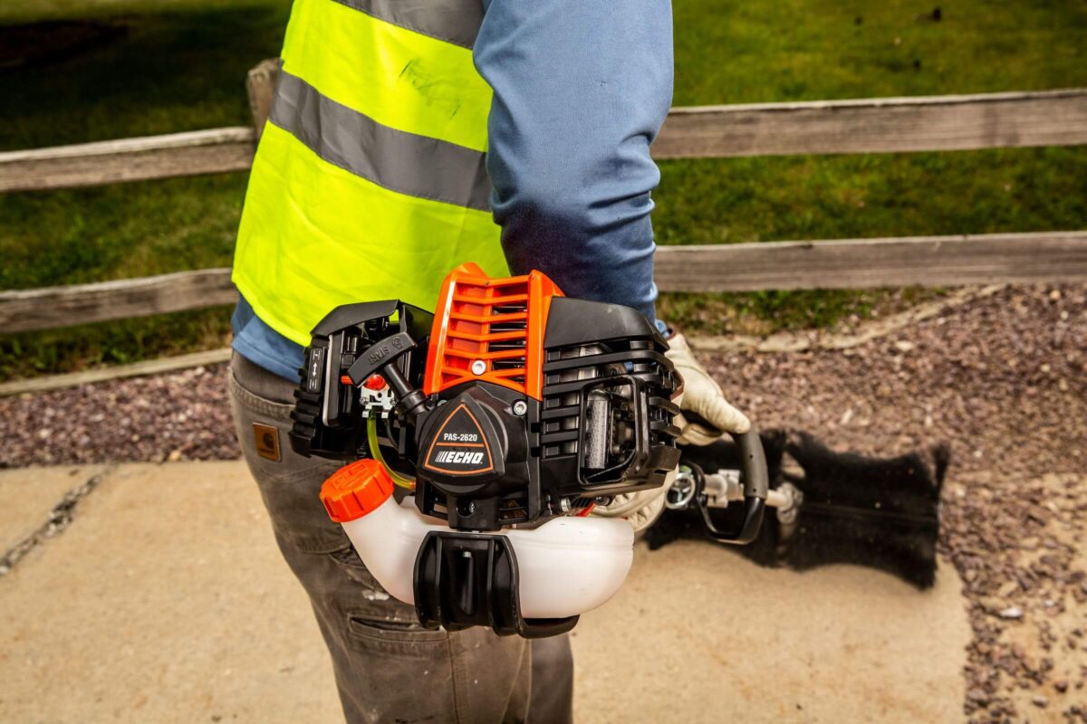 Person wearing a high-visibility vest holds a gas-powered leaf blower. They are standing on a stone path with grass and a wooden fence in the background.