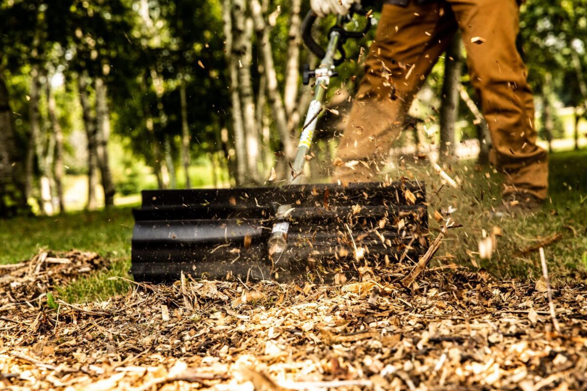 A person operating a wood chipper, shredding branches and producing wood chips. Wood debris is flying in motion in an outdoor setting with trees in the background.