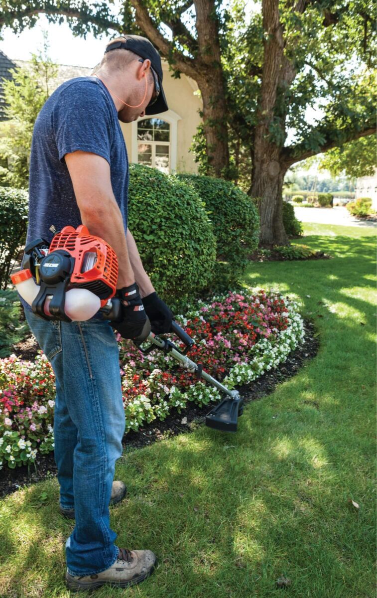 A person in jeans and a blue shirt trims grass edges with a gas-powered trimmer near a flower bed in a garden.