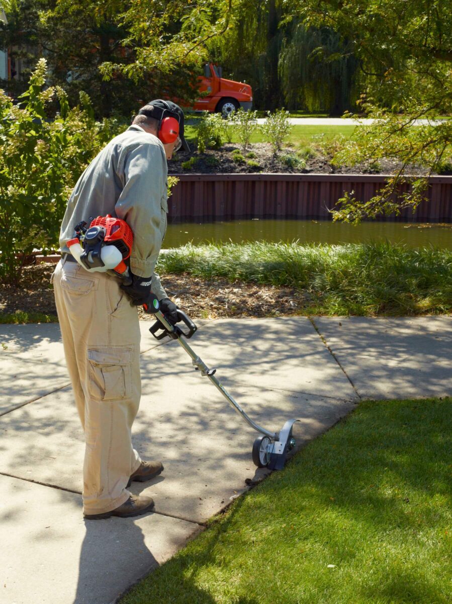 A person wearing protective gear operates a gas-powered grass edger along a sidewalk next to a lawn, with trees and parked car in the background.