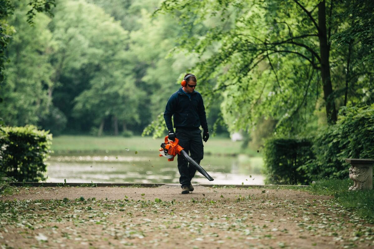 A person using a leaf blower on a dirt path surrounded by trees, wearing protective gear.