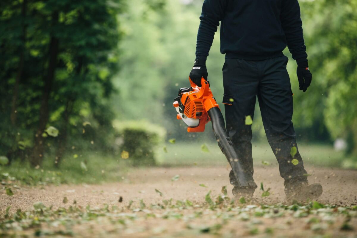 Person in dark clothing uses an orange leaf blower on a dirt path surrounded by greenery.