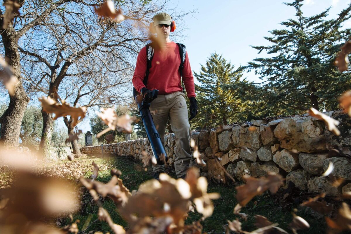 A person wearing protective gear uses a leaf blower to clear fallen leaves along a stone wall path in a park.