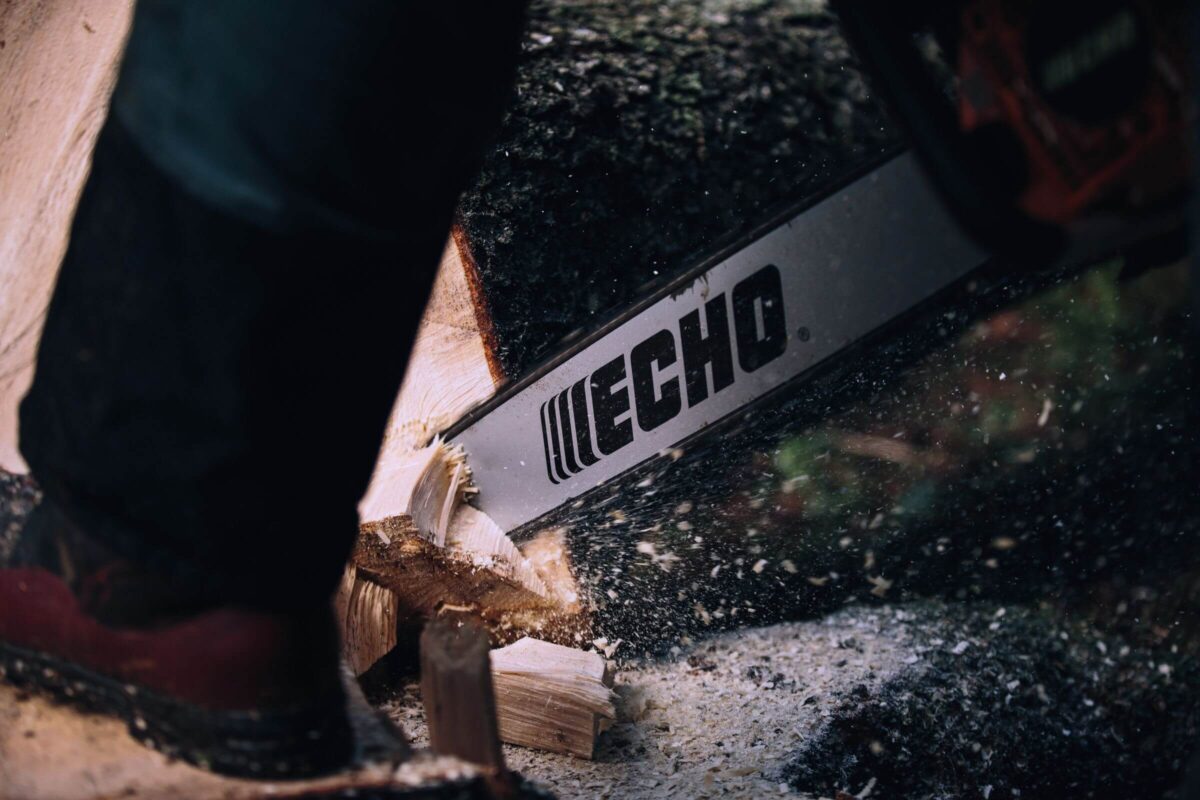 Close-up of a person using an Echo CS-620SX chainsaw to cut through a tree trunk, with wood chips flying around.