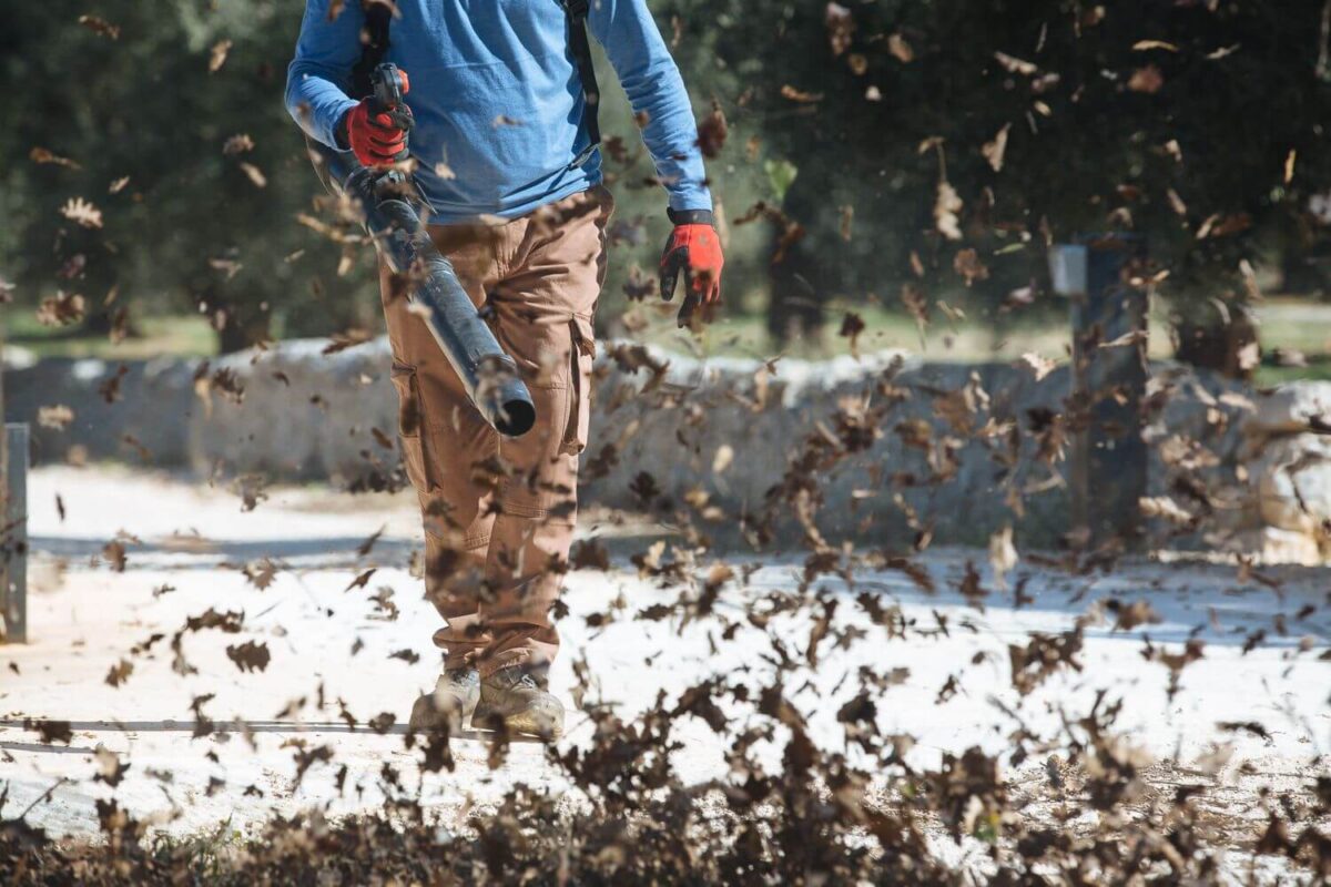 Person using a leaf blower on a paved area, scattering leaves into the air, while wearing a blue shirt, brown pants, and red gloves.