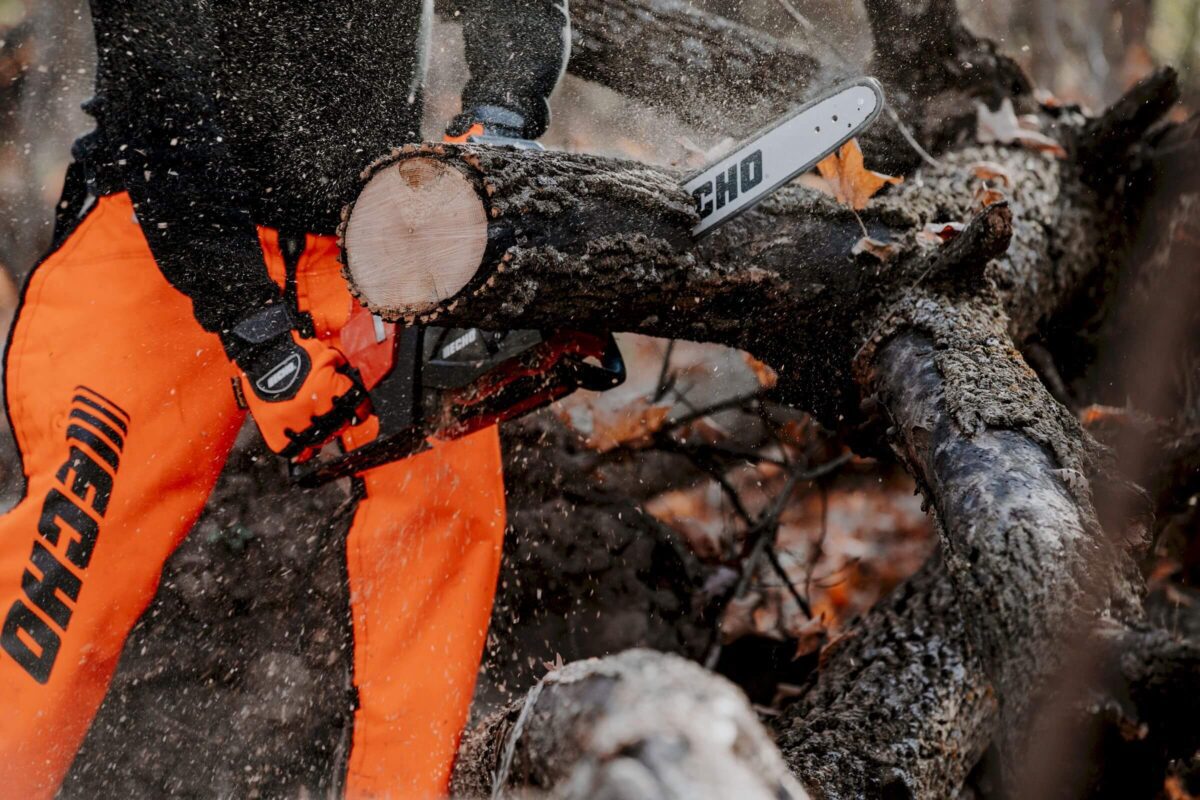 Person in orange safety gear uses a chainsaw to cut a fallen tree branch, with wood chips flying.