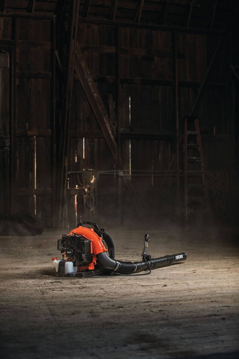 An orange and black leaf blower rests on the dusty floor of a dimly lit barn with wooden walls and a ladder in the background.