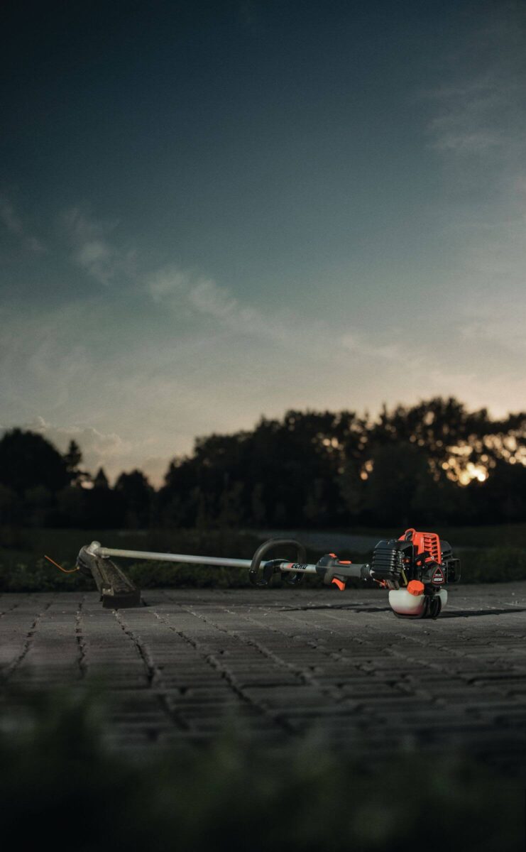 A gas-powered string trimmer with an orange engine rests on a paved surface at dusk, with trees and a cloudy sky in the background.