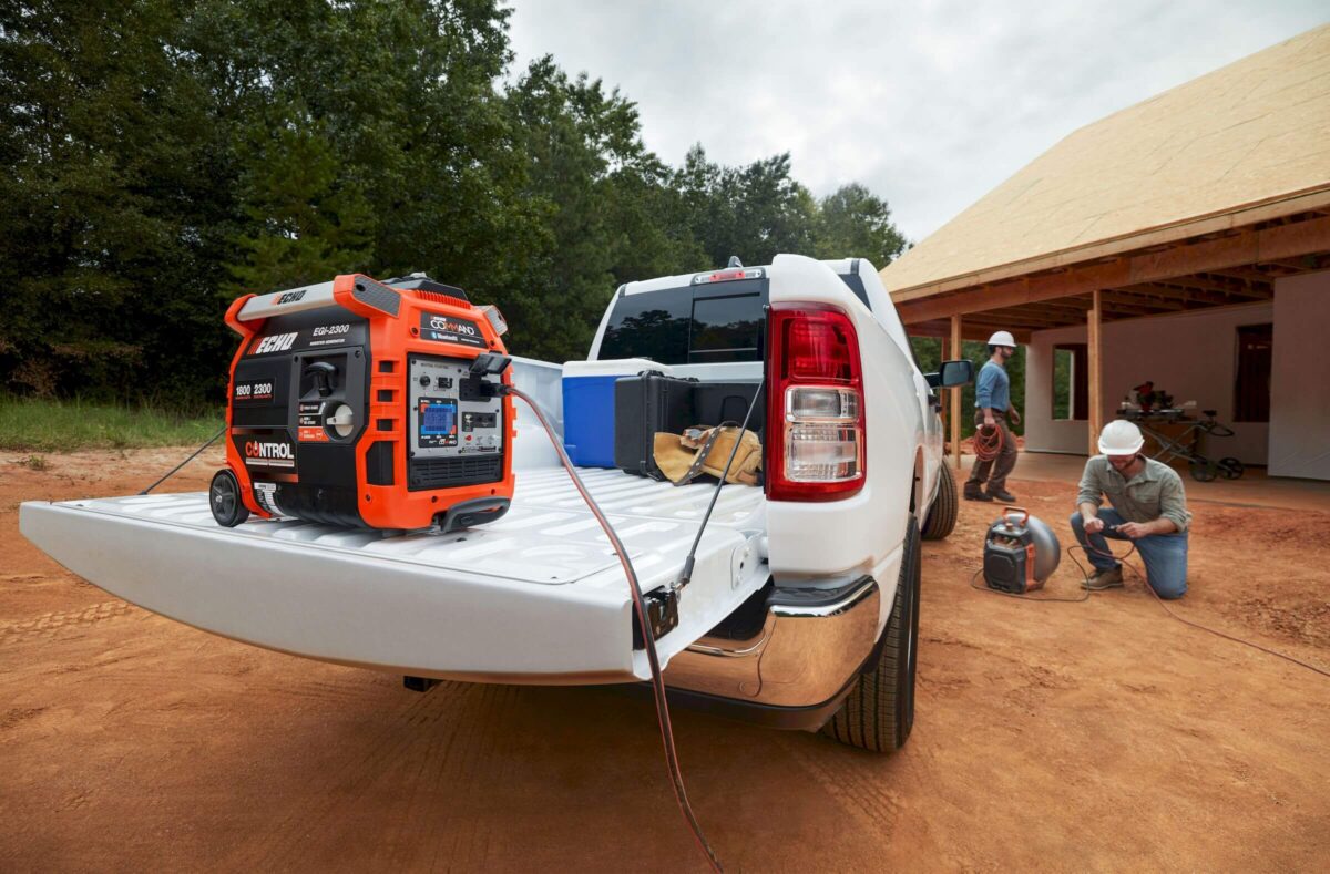 A portable generator is on the tailgate of a pickup truck at a construction site. Two workers in hard hats are setting up equipment alongside.