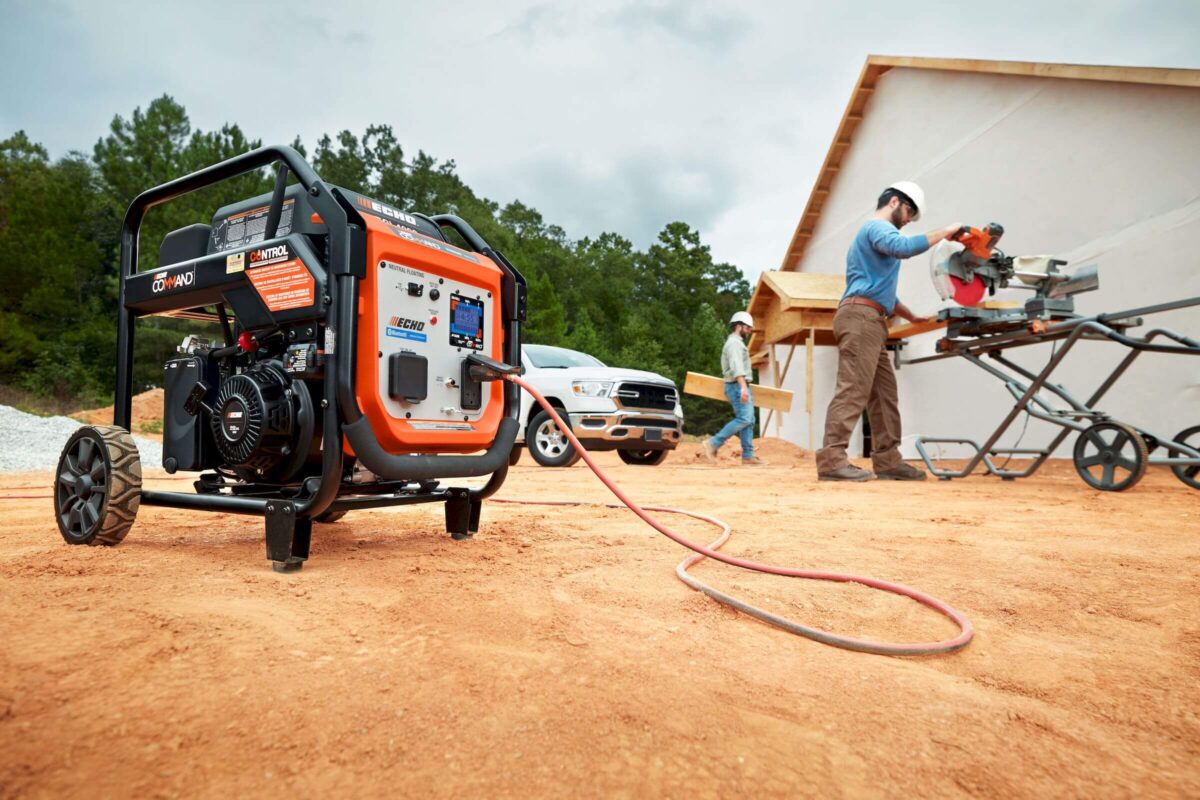 A portable generator powers a saw at a construction site. Two workers in hard hats operate the saw near a wooden structure, with a truck parked nearby.