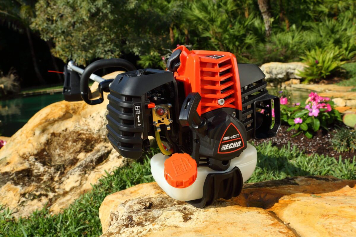 Gas-powered string trimmer with a black and orange body rests on a rock, surrounded by greenery and flowers in a garden setting.