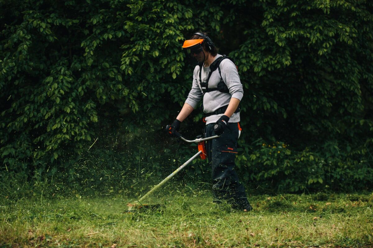 A person wearing safety gear uses a grass trimmer in a lush green garden.